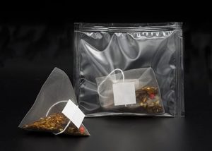 Two transparent pyramid tea bags filled with loose leaf tea, displayed with a resealable plastic pouch, made by Custom Co-Pak, a private label tea packaging company in the USA, on a black background.